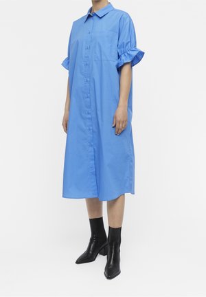 Vestito camicia azzurro chiaro con colletto, chiusura a bottoni, maniche corte a sbuffo e tasca sul petto. La modella indossa stivali neri alla caviglia.