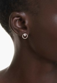 Roséguldfärgade cirkulära örhängen med en central klar sten, omgiven av små klara stenar, uppvisade på mörk hud.