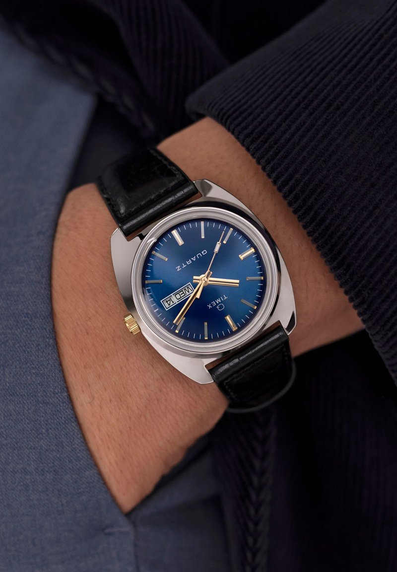 Montre en acier inoxydable argentée avec un cadran bleu, des index des heures dorés et une fonction date. Bracelet en cuir noir avec une texture lisse.