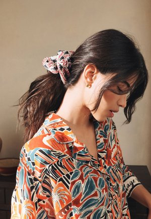 Woman with dark hair tied in a floral scrunchie, wearing colorful patterned shirt, looking down against a plain beige background.