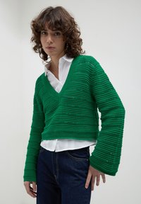 Maglione verde lavorato a maglia con scollo a V e maniche lunghe, caratterizzato da righe orizzontali e un design corto. Indossato sopra una camicia bianca.