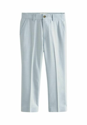 Bambino sorridente con capelli ricci che indossa un abito azzurro chiaro, camicia bianca e scarpe da ginnastica bianche, in piedi davanti a uno sfondo semplice.