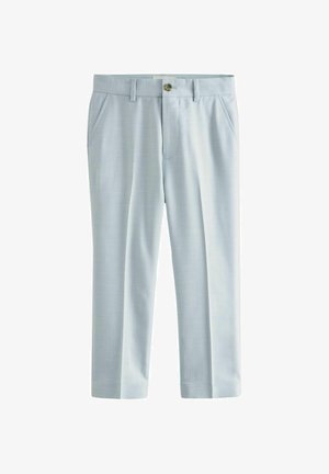 Bambino sorridente con capelli ricci che indossa un abito azzurro chiaro, camicia bianca e scarpe da ginnastica bianche, in piedi davanti a uno sfondo semplice.