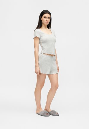 Γυναίκα με μακριά σκούρα μαλλιά, που φοράει ανοιχτό γκρι πλεκτό τοπ με ριμπ ύφανση και κοντά μανίκια, ασορτί σορτς και γκρι χνουδωτά παντόφλες, στέκεται πλάγια.