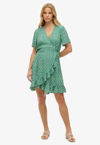 Vestido verde envolvente de tejido ligero con un estampado floral, mangas cortas y evanescentes, y un dobladillo con volantes. Combinado con sandalias beige.