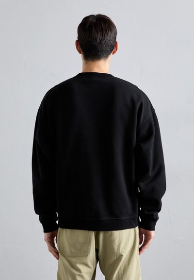 Schwarzes Sweatshirt mit lockerer Passform, Rundhalsausschnitt und überschnittenen Schultern. Der Stoff scheint weich zu sein; getragen mit hellen, locker sitzenden Hosen.