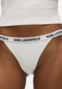 String blanc avec logo noir "KARL LAGERFELD" sur la bande de taille ; matériau lisse, coutures minimales et design taille basse.
