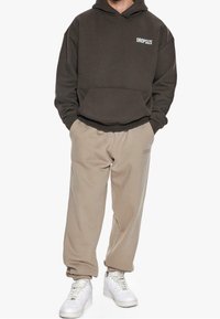 Donkerbruine hoodie met een voorzak en "DROPSIZE" bedrukking, gecombineerd met lichtbeige tapered sweatpants en witte sneakers. Glad textuur.