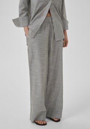 Inimene, kes kannab halli triibulist lahtist pükst paelaga vöökohas ja sobivaid pika varrukaga särke, seisab paljajalu mustade sandaalide peal.