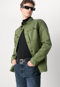 Olivově zelená lehká bunda s dvěma kapsami na hrudi, na knoflíky, nošená přes černý rolák, v kombinaci s tmavě modrými džíny a černým opaskem.