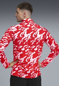 Rode langelijns-shirt met een golvenpatroon in roze, gemaakt van een gladde stof. Het ontwerp bedekt de gehele achterkant. Zwarte broeken zijn zichtbaar.
