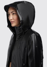 Chaqueta negra impermeable con un acabado brillante, capucha ajustable, cremallera frontal y detalles con cordón. Cuenta con un logo en el pecho.