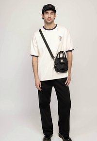 White t-shirt with navy trim, black cargo pants, and a small black bag with a logo, completed with a black cap. Model standing against a neutral background.
