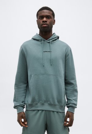 Mann trägt einen gedämpften grünen Hoodie und passende Shorts, steht vor einem einfachen hellen Hintergrund und blickt neutral nach vorne.