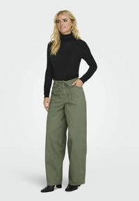 Schwarzer Rollkragenpullover kombiniert mit hochtaillierten, locker sitzenden, olivgrünen Hosen mit gebundenem Taillenbund und Taschen. Schwarze Absatzschuhe.