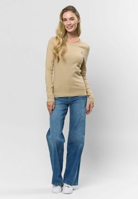 Hosszú ujjú bézs bordázott felső, világoskék, trapéz farmernadrággal és fehér sportcipővel párosítva. A felső kerek nyakkal és testhez simuló fazonnal rendelkezik.