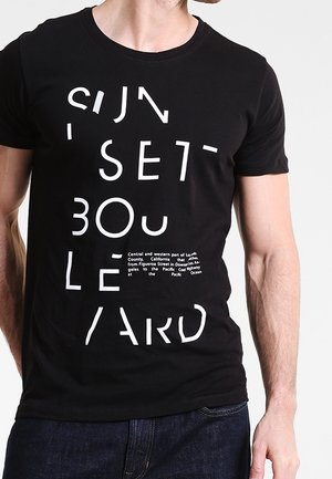 Mand iført en sort t-shirt med store, spredte hvide bogstaver, der læser "SUNSET BOULEVARD", og lille hvid tekst nedenunder.