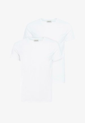 Junger Mann mit lockigem Haar, der ein schlichtes weißes V-Ausschnitt-T-Shirt, graue plissierte Hosen und weiße Sneaker trägt und vor einem weißen Hintergrund steht.