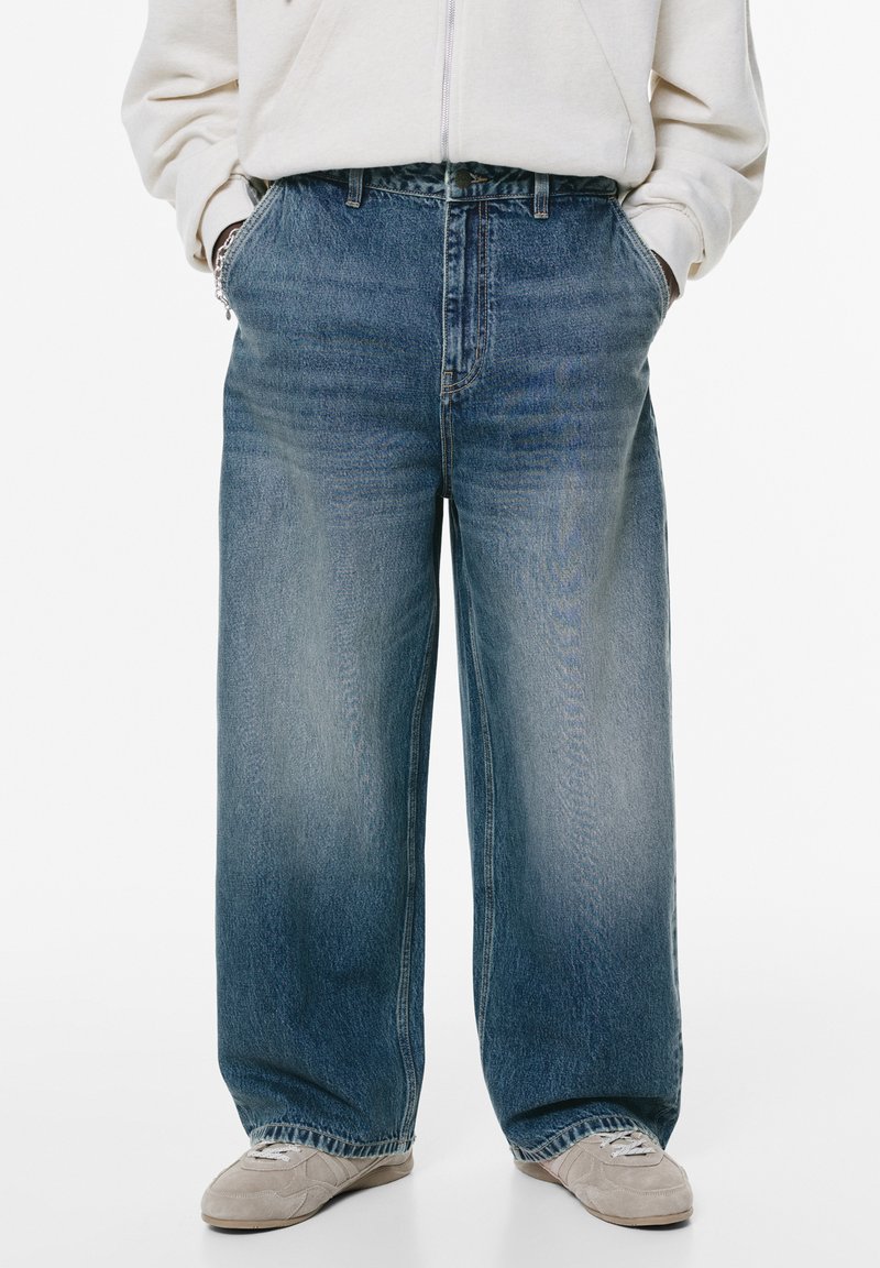 Jeans in denim blu a gamba larga con una texture sbiadita, vita alta e design tradizionale a cinque tasche. Abbinati a un maglione grigio chiaro.