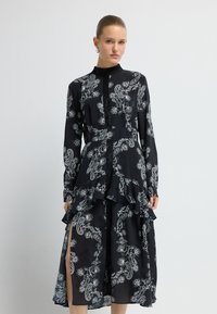 Kobieta ubrana w czarną sukienkę midi z długim rękawem, ozdobioną białymi kwiatowymi wzorami paisley i falbanami, stojąca na tle jednolitego, białego tła.