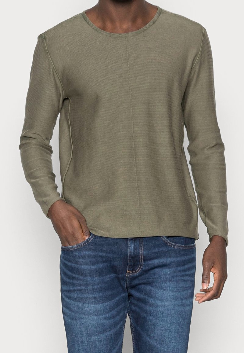 Chemise à manches longues vert olive avec une texture côtelée, un col rond et des détails de coutures visibles. Portée avec un jean bleu.