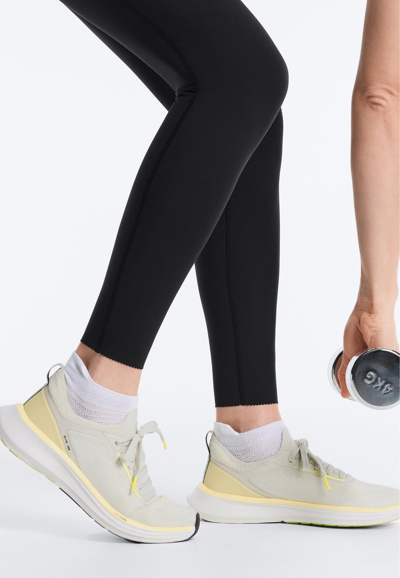 Personne en leggings noirs et baskets jaune clair tenant un haltère de 4 kg, en position de fitness ou d'exercice.