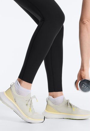 Person i sorte leggings og lysegule sneakers holder en 4 kg håndvægt, klar i en fitness- eller træningsstilling.