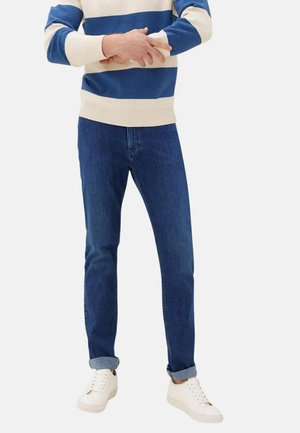 Mężczyzna w niebiesko-białym swetrze w paski, ciemnoniebieskich podwiniętych dżinsach i białych trampkach, stoi z założonymi na siebie ramionami.