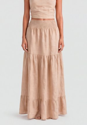 Kobieta w beżowej warstwowej maksi spódnicy z marszczoną talią i dopasowanym krótkim, bezrękawnym topem, stojąca na jednolitym tle.