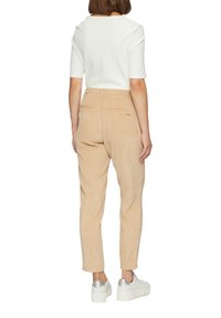 Beige Cord-Hosen mit schmalem Schnitt, ausgestattet mit Seitentaschen, Gesäßtaschen und einem elastischen Bund. Getragen mit einem weißen gerippten Oberteil und Sneakers.