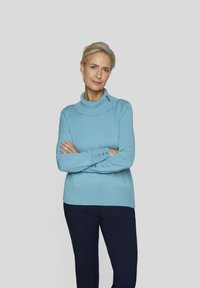 Helles blaues Strickpullover mit hohem Kragen und Knopfdetails an den Ärmeln. Kombiniert mit dunkelblauen Hosen, die eine glatte Textur aufweisen.