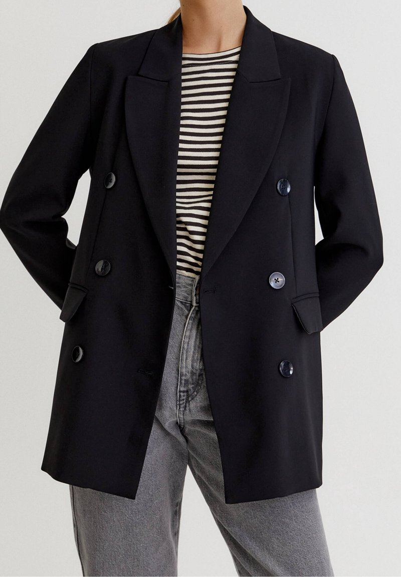 Une personne porte un blazer noir croisé par-dessus une chemise rayée noir et blanc avec un jean taille haute gris clair, les mains derrière le dos.