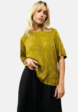 Olijfgroene blouse met korte mouwen en een bladerenpatroon, gemaakt van zachte stof, met een ontspannen pasvorm en een ronde zoom, gecombineerd met een zwarte rok.