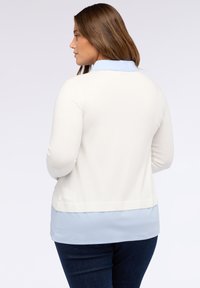 Femeie cu păr brunet îmbrăcată într-un pulover crem peste o cămașă albastră deschis cu guler și blugi negri, întoarsă lateral cu un fundal simplu.