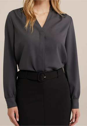 Mujer vestida con una blusa de manga larga gris oscuro metida en una falda negra de cintura alta con un cinturón negro.