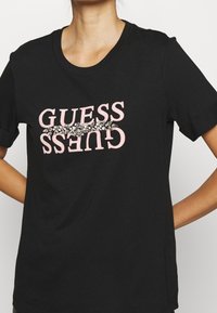 Černé bavlněné tričko s růžovým logem "GUESS", ozdobené dekorativními kamínky podél písmen. Krátké rukávy a klasický kulatý výstřih.