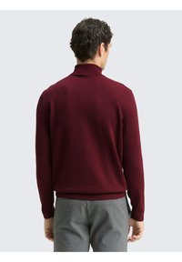 Bordeauxroter, gestrickter Rollkragenpullover mit gerippten Bündchen und Saum, kombiniert mit grauer Hose. Glatte Oberfläche, figurbetontes Design.