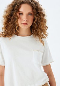 Camiseta de algodón blanca con mangas cortas, escote redondo y un bolsillo en el pecho. Presenta costuras en contraste a lo largo de los bordes. Textura suave.