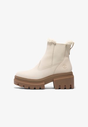 Botas de tobillo beige con suela de goma marrón chunky, borde de piel suave en la parte superior y paneles elásticos en los laterales; presenta un logo en el talón.