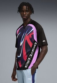 Νέος άνδρας με πλεγμένα μαλλιά που φοράει αθλητική μπλούζα σε μαύρο, ροζ και κόκκινο με αφηρημένο σχέδιο και ανοιχτόχρωμο μπλε φόρμα Puma.