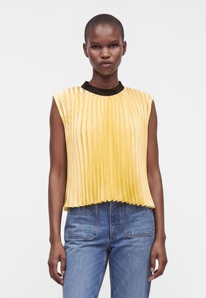 Femme aux cheveux courts portant un haut plissé jaune sans manches avec un col noir et un jean bleu, debout devant un fond blanc uni.