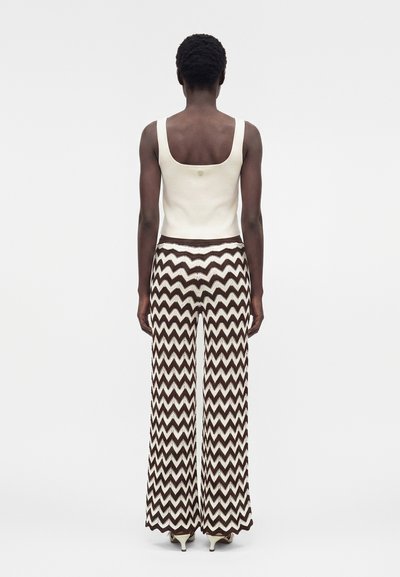 Άτομο στέκεται με την πλάτη, φοράει κρεμ τοπ χωρίς μανίκια και καφέ-λευκά φαρδιά παντελόνια με ζιγκ-ζαγκ μοτίβο και τακούνια.