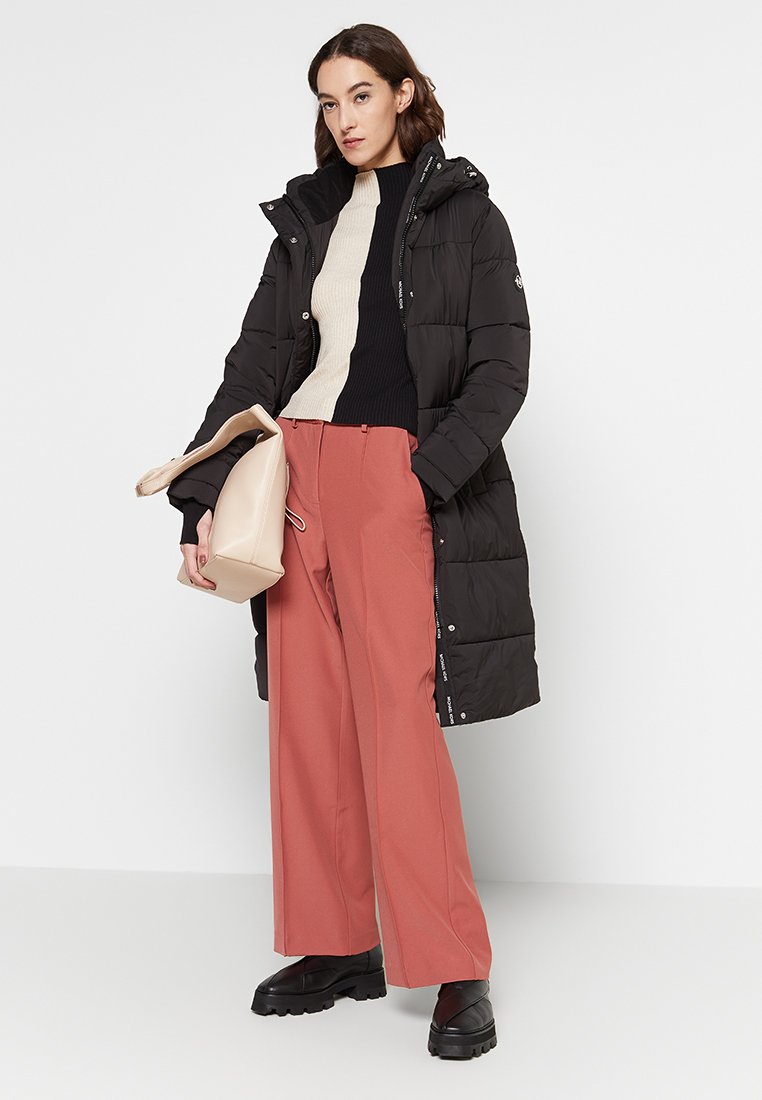 Schwarze Daunenjacke mit Kapuze, lange Form, getragen über einem creme- und schwarz gestreiften Pullover, kombiniert mit pfirsichfarbenen weiten Hosen und schwarzen Stiefeln.