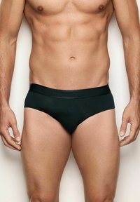 Torso maschile e parte superiore delle gambe che indossa slip neri aderenti con una cintura con marchio, in piedi su uno sfondo chiaro e uniforme.