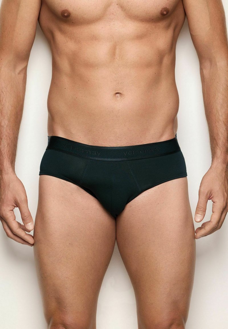 Torso maschile e parte superiore delle gambe che indossa slip neri aderenti con una cintura con marchio, in piedi su uno sfondo chiaro e uniforme.