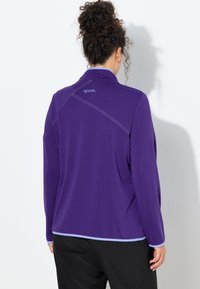 Fioletowa bluzka z długim rękawem, z wysokim kołnierzem, kontrastowym przeszyciem i logo na plecach. Gładka faktura i luźny krój.