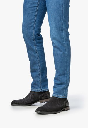 Botas Chelsea negras con acabado texturizado, combinadas con jeans de mezclilla azul ajustados, que tienen una pierna estrecha y un ligero desvanecimiento cerca de los tobillos.