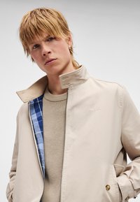 Μπεζ σακάκι με ψηλό γιακά, με μπλε καρό φόδρα. Φορεμένο πάνω από μια υφασμάτινη μπεζ ζακέτα, έχει φερμουάρ και διακοσμητικά κουμπιά.