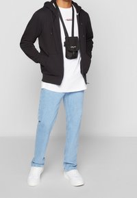 Svart zip-up hoodie, vit t-shirt, ljusblå jeans och vita sneakers. En svart crossbody-väska hänger vertikalt och har en logotyp.