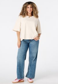 Vrouw met krullend haar die een beige oversized shirt, blauwe spijkerbroek, roze sneakers en gouden sieraden draagt, staat zelfverzekerd met één hand in de zak.